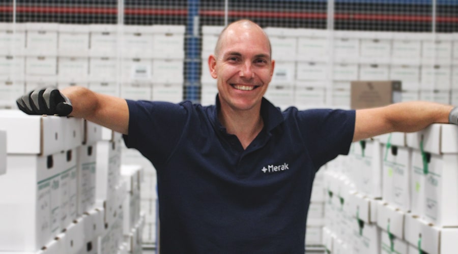 Un homme souriant portant un polo de travail bleu Merak s'appuie détendu contre des piles de boîtes dans un entrepôt, reflétant un environnement de travail organisé et professionnel.