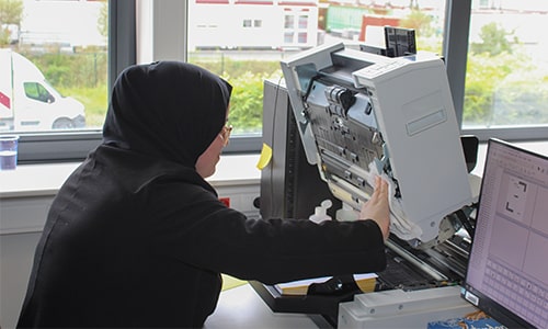 Fatima, une employée portant un voile, utilise un grand canner dans un bureau lumineux. Elle travaille minutieusement en vérifiant la machine.