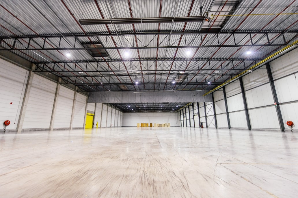 Smiling man in gray sweater walking through a warehouse filled with wooden crates.